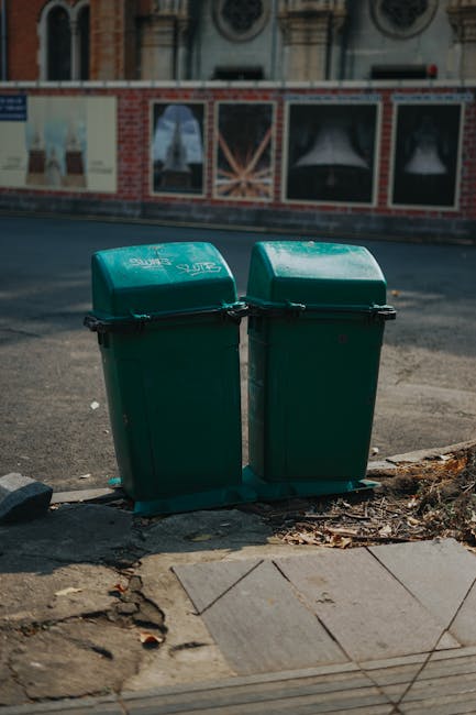 Two green wheeled rubbish bins with hinged lids are positioned on a concrete sidewalk curb in an urban environment. The bins are made of plastic with a textured surface and are branded with a white logo and text. They are placed side by side, slightly leaning toward each other, and are secured with black straps around the middle. The pavement around the bins features scattered leaves, small debris, and some cracked tiles. Behind the bins, a street runs parallel, with a building facade visible in the background that includes a row of windows with decorative frames and brickwork. The scene is lit by daylight, with natural shadows cast across the sidewalk and street surface. The presence of these bins in an accessible outdoor location suggests they are designated for waste disposal or collection, supported by waste management services like Waste Disposal Highbury specializing in rubbish removal, including alternative collection methods outside of local council schemes.