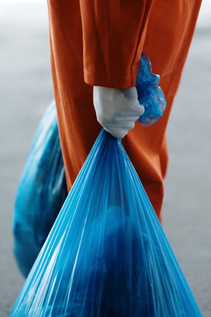 Close-up view of a person dressed in orange overalls wearing white gloves, holding a tightly knotted blue plastic rubbish bag by its gathered top, with their hand positioned near the bag’s neck. The bag appears full and made of thin, semi-translucent plastic material, showing a hint of its contents. The background is blurred, suggesting an outdoor setting possibly at a waste collection point or disposal area. The scene emphasizes responsible waste handling and disposal, aligning with the context of rubbish removal services provided by Waste Disposal Highbury, including private collection and on-site clearance. The lighting is natural but diffuse, with no strong shadows, highlighting the texture of the glove, the sheen of the plastic bag, and the vibrant colour contrast between the orange attire and blue rubbish bag. Overall, this image exemplifies the process of safe and professional waste disposal, supporting the primary focus on rubbish removal in an urban environment.