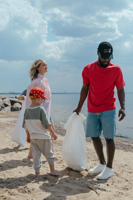 A group of three individuals, including a man and two children, are standing on a sandy beach near the water's edge during the daytime. The man, dressed in a red t-shirt, light blue denim shorts, and a black cap, is holding a large, clear plastic rubbish bag and appears to be collecting litter. The two children, a girl with curly blonde hair wearing a light pink jacket and an older boy in a grey t-shirt and grey trousers with a red cap featuring yellow and white patterns, are also engaged in the activity, with one of them holding a bag. In the background, the sky is partly cloudy with patches of blue, and the shoreline is lined with rocks. This scene reflects a community or family effort in outdoor waste management and environmental care, aligning with private rubbish removal and on-site clearance services, potentially offered by Waste Disposal Highbury.