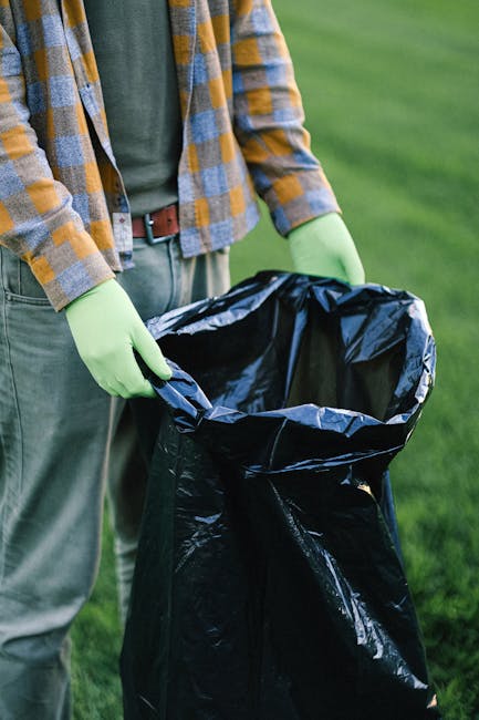 Close-up view of a person dressed in orange overalls wearing white gloves, holding a tightly knotted blue plastic rubbish bag by its gathered top, with their hand positioned near the bag’s neck. The bag appears full and made of thin, semi-translucent plastic material, showing a hint of its contents. The background is blurred, suggesting an outdoor setting possibly at a waste collection point or disposal area. The scene emphasizes responsible waste handling and disposal, aligning with the context of rubbish removal services provided by Waste Disposal Highbury, including private collection and on-site clearance. The lighting is natural but diffuse, with no strong shadows, highlighting the texture of the glove, the sheen of the plastic bag, and the vibrant colour contrast between the orange attire and blue rubbish bag. Overall, this image exemplifies the process of safe and professional waste disposal, supporting the primary focus on rubbish removal in an urban environment.