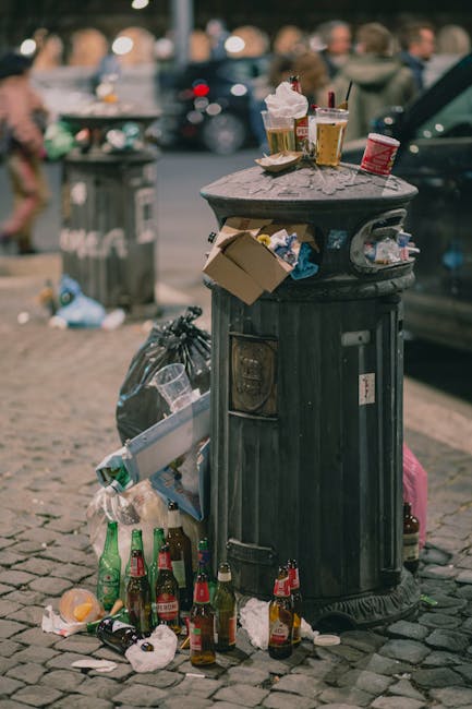 A black, cylindrical public litter bin made of metal with vertical ridges along its surface, placed on a cobblestone pavement in an urban area. The top of the bin is covered with scattered discarded waste, including takeaway coffee cups with plastic lids, crumpled paper, and food packaging. Surrounding the base are several empty glass bottles, some with labels visible, alongside discarded paper and plastic waste, indicating recent outdoor consumption and littering. In the background, blurred figures and parked cars suggest a busy street scene. The scene illustrates typical urban waste accumulation that might require collection or clearance services, such as those offered by Waste Disposal Highbury for managing roadside rubbish and maintaining cleanliness in residential or commercial areas through private waste handling solutions.