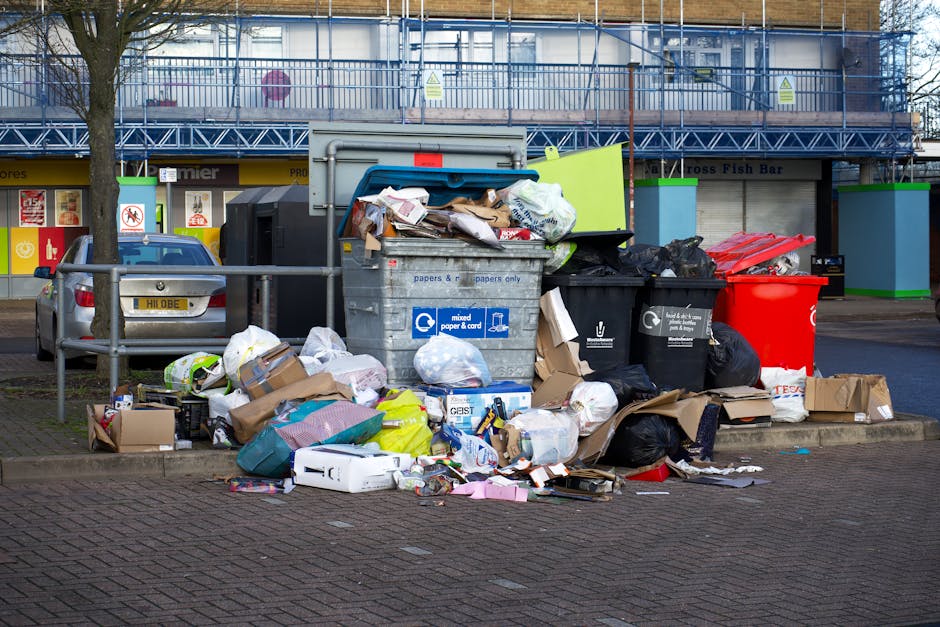 A large collection of mixed household waste and recycling within and around multiple rubbish bins situated on a paved urban street; the central metal bin is overflowing with paper, cardboard boxes, and plastic packaging, some of which are spilling onto the ground. Several black and red plastic rubbish bags are placed beside the bins, with additional discarded items including flattened cardboard, plastic containers, and packaging material scattered at the base. The background features a commercial building with a blue scaffold, green fencing, and signage for shops, indicating the waste is positioned near retail outlets or a shopping area. A parked grey car is partially visible to the left, and a metal railing separates the waste area from the street. The scene is set during daylight hours, with natural light illuminating the cluttered environment, reflecting a typical instance where independent rubbish collection or private waste disposal services, such as those provided by Waste Disposal Highbury, might be involved in clearing excess waste from residential or commercial properties.