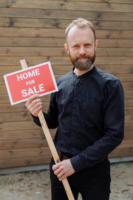 A man with a beard and short hair, wearing a dark long-sleeved shirt, stands outdoors in front of a wooden fence. He holds a small wooden signpost with a rectangular, bright red and white sign that reads 'HOME FOR SALE'. The sign is positioned at chest height and slightly tilted, supported by the man's right hand. The ground appears to be a concrete or paved surface with some dirt patches. The scene suggests a real estate context, but in relation to rubbish removal services, it illustrates a typical setting where private property is being prepared for sale or rent, potentially involving clearance or waste management tasks. The natural lighting indicates a clear day, highlighting the textures of the man's clothing and the wooden fence behind him.