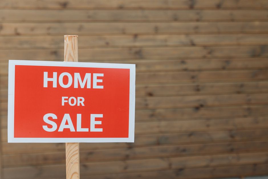 A rectangular sign with a bold red background and white uppercase text displaying the words 'HOME FOR SALE' is mounted on a natural wooden stake. The stake is inserted into the ground, partially visible at the top of the sign, and has a smooth, light-colored finish. The sign is positioned in front of a wooden fence made of horizontal planks, which have a weathered appearance with visible grain and knots, in varying shades of brown. The background fence extends across the entire image, creating a neutral setting that emphasizes the sign. The scene is evenly lit, with no strong shadows or highlights, suggesting daylight conditions. The image captures a typical context of property marketing, often associated with estate agencies or private sellers, indirectly related to the management and disposal of waste and rubbish associated with house sales or renovations, which could involve services from Waste Disposal Highbury in managing clearance or waste removal on-site.