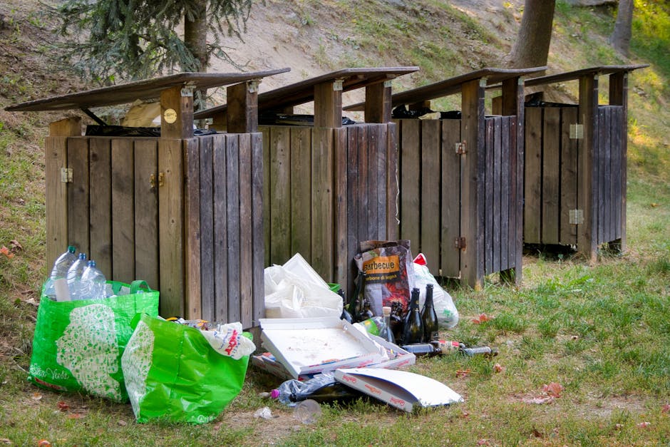 A row of four wooden outdoor waste bins with slanted roofs, situated on a grassy area near a gentle slope with trees in the background. The bins are made of weathered dark brown wood, each with metal hinges and locks, and are positioned side by side along a narrow strip of ground. In front of the bins, various discarded items are scattered on the grass, including a large green reusable shopping bag filled with plastic bottles, a white cardboard pizza box, several empty glass bottles, and plastic bags containing other waste. Some of the bottles appear to be used for beverages, while others are clearly waste items. The scene is outdoors with natural lighting, and the environment suggests a public park or garden area where waste has been left nearby, reflecting an on-site or independent waste disposal scenario handled by Waste Disposal Highbury within a rubbish management context.