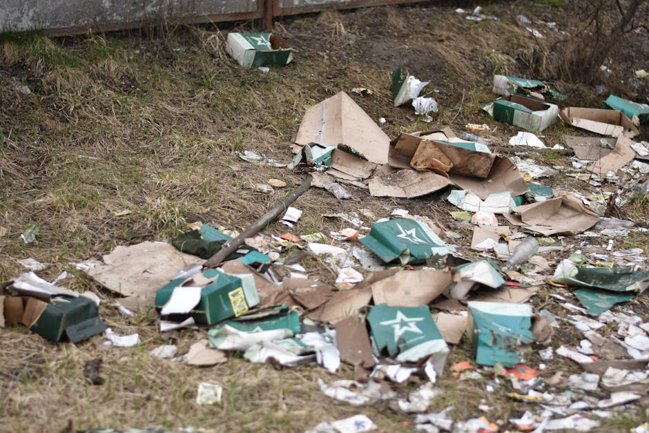 A row of four wooden outdoor waste bins with slanted roofs, situated on a grassy area near a gentle slope with trees in the background. The bins are made of weathered dark brown wood, each with metal hinges and locks, and are positioned side by side along a narrow strip of ground. In front of the bins, various discarded items are scattered on the grass, including a large green reusable shopping bag filled with plastic bottles, a white cardboard pizza box, several empty glass bottles, and plastic bags containing other waste. Some of the bottles appear to be used for beverages, while others are clearly waste items. The scene is outdoors with natural lighting, and the environment suggests a public park or garden area where waste has been left nearby, reflecting an on-site or independent waste disposal scenario handled by Waste Disposal Highbury within a rubbish management context.