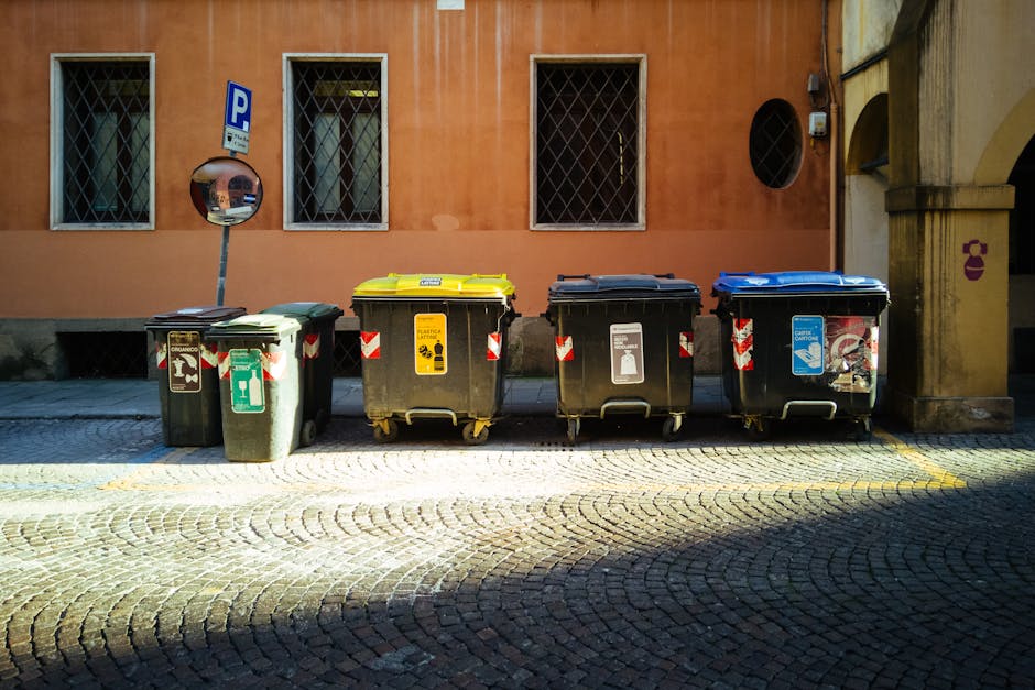 A row of four large wheeled rubbish bins positioned on a cobblestone street in front of a salmon-colored building with three windows featuring diamond-patterned metal security grills. The first bin on the left is dark green with a rounded lid and a recycling symbol, and beside it is a black bin with a yellow lid designated for general waste, displaying a hazardous waste warning label. The third bin is black with a dark gray lid, with a white label indicating recyclable waste, and the fourth bin, also black with a blue lid, bears a label for recyclable materials. Behind the bins, a reflective road sign indicating parking is mounted on a metal pole, casting slight shadows on the ground. The environment appears to be a quiet urban setting possibly within a designated waste disposal zone, reflecting typical private rubbish handling with multiple waste types stored for collection by waste management services like Waste Disposal Highbury, aligning with the context of alternative waste handling options.