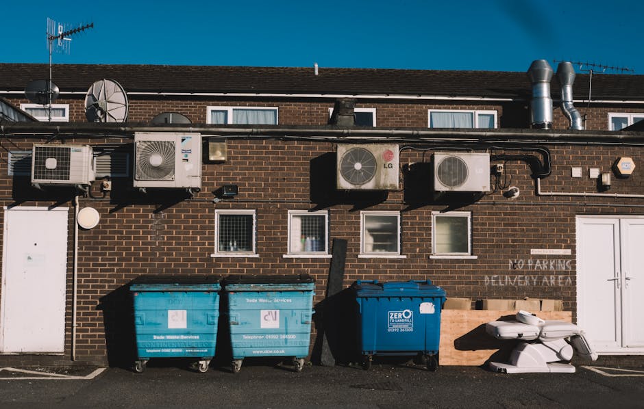 The image depicts a brown brick building with three small, rectangular windows on the upper level and two larger doors on the ground level. Mounted on the exterior wall above the ground, there are several air conditioning units and ventilation pipes, with some pipes stainless steel and others black, running horizontally across the wall. To the left, a satellite dish is attached to the wall, and there are antennas visible atop the building's roof. In front of the building, on a paved surface, there are three large wheelie bins made of plastic; two are bright blue, designated for waste collection, and one is darker blue with a white logo indicating it is for waste disposal services. The bins are closed, with lids secured, and are positioned side by side. To the right of the bins, there are two white plastic items that resemble discarded or abandoned objects, possibly parts of furniture or construction debris. The background features a clear blue sky, suggesting a bright, sunny day. The scene appears to be an urban setting suitable for private or alternative waste disposal, consistent with rubbish removal or on-site clearance services provided by Waste Disposal Highbury.