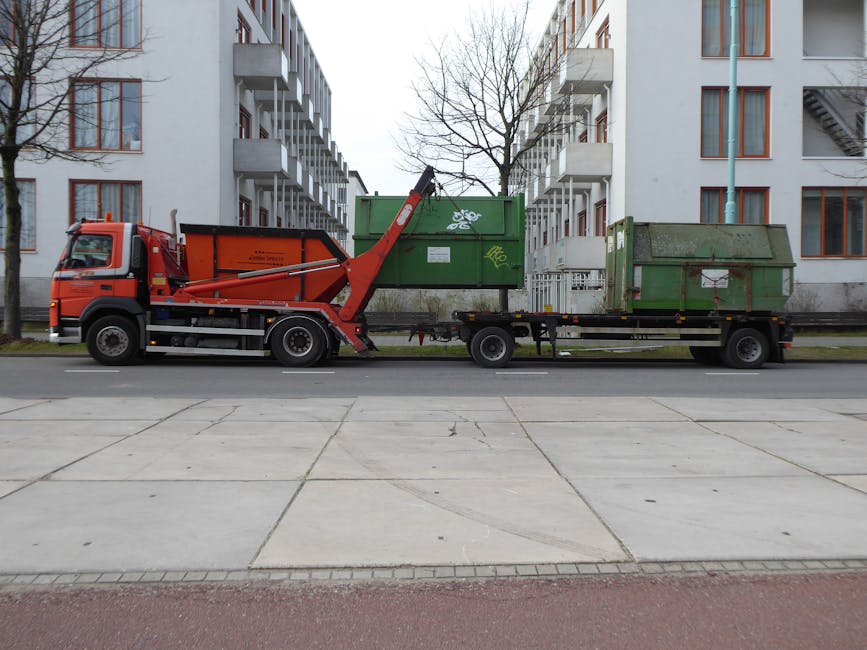 A flatbed truck parked on a paved street in front of a modern, white residential building with multiple stories and balconies. The truck features an orange and black grab lift mechanism mounted on the driver's side, used for offloading and handling waste containers. On the truck's trailer, there are two large, green metal waste bins; one with a floral graffiti design and the other with rust and dirt marks. The bins are situated side by side, with the larger one positioned slightly behind the smaller, both showing signs of wear and weathering, including patches of moss or grime. Surrounding the scene, there are leafless trees and a metal fence separating the street from the building's premises. The lighting is natural and diffuse, typical of an overcast day. This image illustrates private waste collection services involving on-site container handling, reflecting an alternative approach to traditional rubbish disposal.