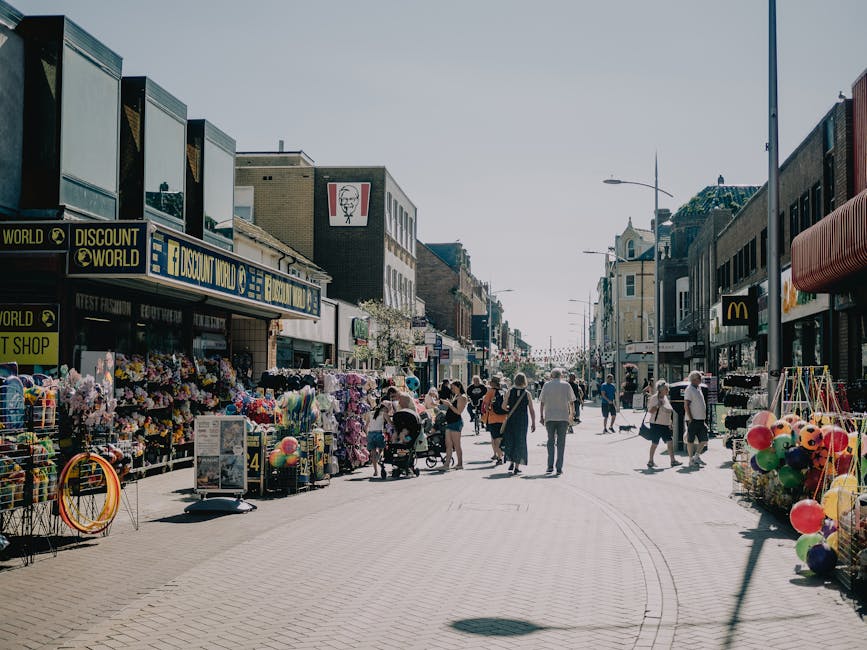 A busy street scene in a high street shopping area with pedestrians walking along the paved walkway. On the left side, there are several retail stores, including a shop with a large sign reading 'Discount World' and displays of various goods such as flowers, balloons, and toys arranged outside. The storefronts are multi-story with modern facades, featuring large windows, signage, and some decorative elements. On the right side, more shops are visible, including a McDonald's outlet, with some outdoor displays also present. Street lamps and a clear sky overhead suggest daytime with bright natural lighting. People of different ages, some pushing strollers or carrying shopping bags, are engaged in walking, shopping, and casual conversation, contributing to an active, commercial atmosphere. The scene depicts an urban retail environment with a mix of commercial activity and pedestrian movement, typical of a high street, and highlights the importance of efficient rubbish collection services such as those provided by Waste Disposal Highbury for maintaining cleanliness in busy shopping districts.