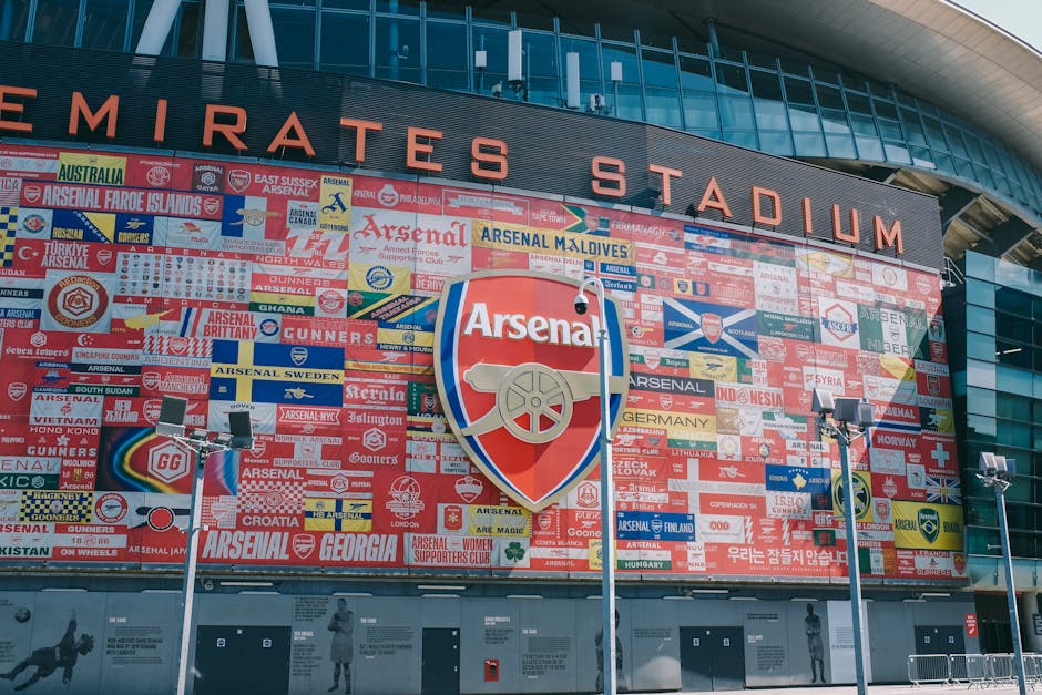 A large exterior wall of Emirates Stadium, home to Arsenal Football Club, features a prominent installation of numerous small, colorful banners displaying the flags and emblems of various countries and regions, arranged in a mosaic pattern. In the center, the Arsenal shield logo, which includes a red background with a gold cannon, is mounted on the wall. The building's modern architecture is highlighted by curved glass panels and a sleek metal facade, with the stadium's name displayed in large, bold, red letters across the top of the structure. In front of the wall, there are three metallic lampposts equipped with floodlights, pointing towards the display to illuminate it at night. The environment appears to be outdoors, on a paved area with no visible pedestrians or vehicles near the scene, emphasizing the detailed mural as a focal point in the stadium's exterior setting.