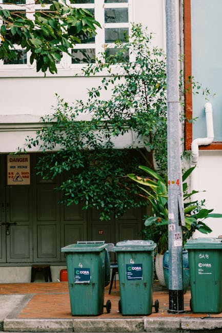 In the image, three green wheeled rubbish bins with black wheels and blue labels are positioned on a paved sidewalk in front of a street-side building. The bins are arranged side by side with their lids closed, and they appear to be made of plastic with a textured surface. Behind the bins, a small green shrub with leafy branches partially obscures a dark green external storage compartment or utility box attached to the building, which features a warning sign with hazard symbols, indicating electrical or other dangers. To the right of the bins, there is a tall, slim metal pole with a grey finish and a vertical white pipe attached, and a smaller orange pipe or conduit nearby, possibly part of the building's drainage or utility system. The backdrop includes the building’s white exterior wall, a window with multiple small panes above, and a section of orange brickwork to the top right, suggesting a typical urban setting. The scene suggests a location where private waste collection or on-site rubbish disposal might be managed independently, supported by Waste Disposal Highbury's services.