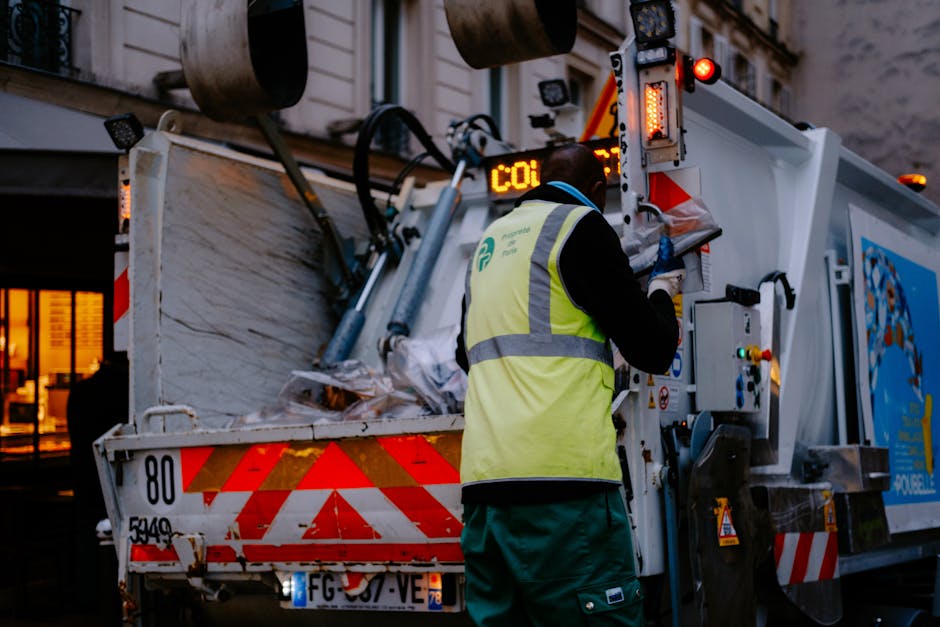 A waste collection worker wearing a high-visibility yellow vest with reflective stripes is operating a large, white waste disposal truck on a city street during dusk or early evening. The truck's rear compartment is open, revealing a load of mixed rubbish, including cardboard and plastic bags, positioned on a metal platform with red and white reflective safety markings. The worker's back is to the camera as they interact with controls or a display panel situated on the side of the vehicle, which features various buttons, levers, and warning lights. The background shows a building with illuminated windows, indicating an urban environment, while the overall scene is dimly lit with some ambient street lighting. The image correlates with private waste management services aimed at alternative or independent waste collection, supporting rubbish removal for flats or multi-occupancy buildings in Highbury, as managed by Waste Disposal Highbury.