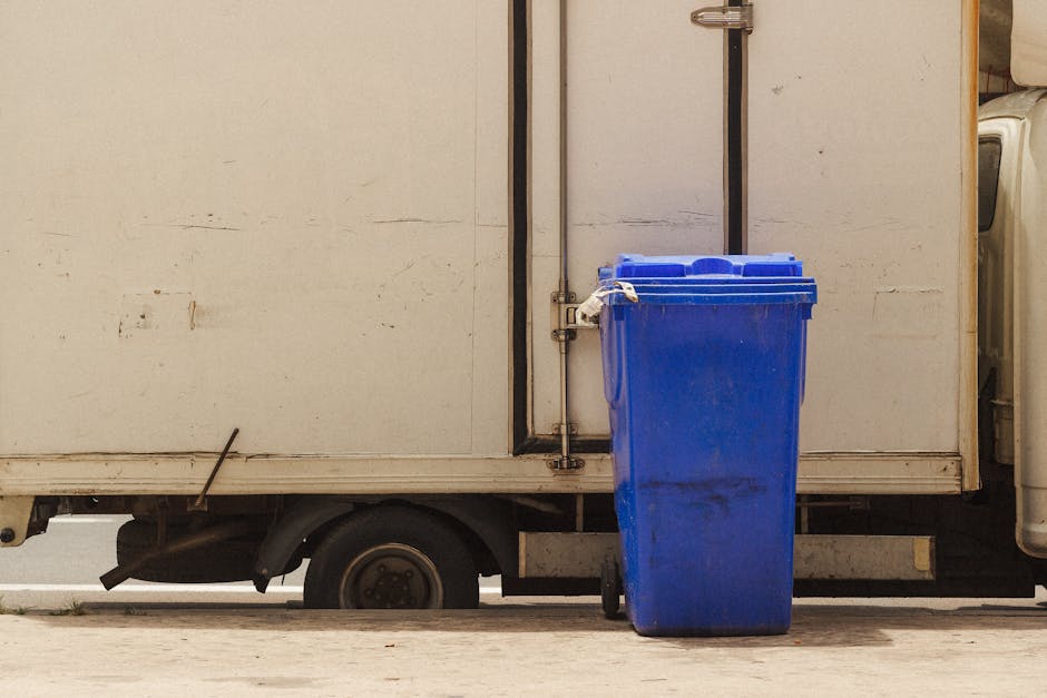 In the image, three green wheeled rubbish bins with black wheels and blue labels are positioned on a paved sidewalk in front of a street-side building. The bins are arranged side by side with their lids closed, and they appear to be made of plastic with a textured surface. Behind the bins, a small green shrub with leafy branches partially obscures a dark green external storage compartment or utility box attached to the building, which features a warning sign with hazard symbols, indicating electrical or other dangers. To the right of the bins, there is a tall, slim metal pole with a grey finish and a vertical white pipe attached, and a smaller orange pipe or conduit nearby, possibly part of the building's drainage or utility system. The backdrop includes the building’s white exterior wall, a window with multiple small panes above, and a section of orange brickwork to the top right, suggesting a typical urban setting. The scene suggests a location where private waste collection or on-site rubbish disposal might be managed independently, supported by Waste Disposal Highbury's services.