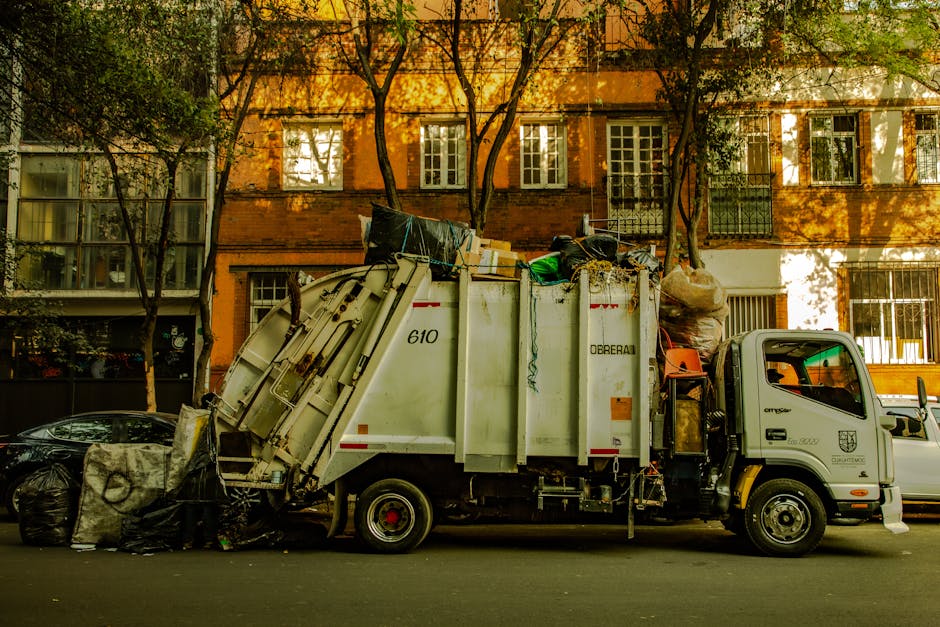 A white garbage collection truck parked on a city street in front of a multi-storey brick building with large windows. The truck is loaded with various waste materials, including black trash bags, cardboard boxes, and other refuse piled on top of the vehicle's open container. The side of the truck features identification numbers and branding, with some debris hanging over the rear edge. Adjacent to the truck, on the pavement, there are additional black trash bags and loose waste. The background includes several tall trees with green foliage casting shadows on the building, and a few parked cars are visible along the street. The scene appears to depict a routine rubbish collection, possibly involving private or independent waste disposal, aligned with waste removal services provided by Waste Disposal Highbury. The overall environment suggests an urban area with organized waste management handling large amounts of refuse for disposal or recycling.